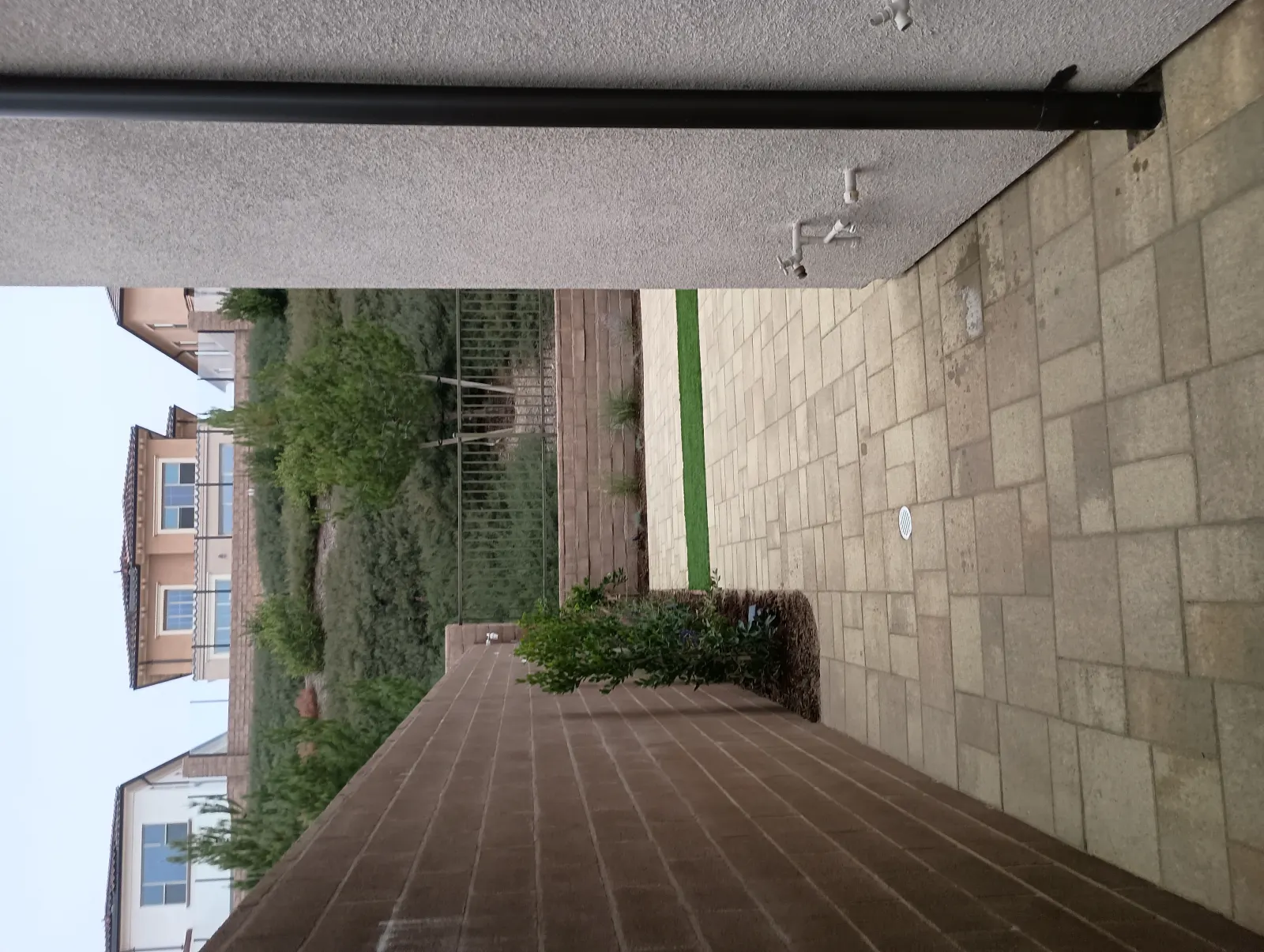 Sideways photo showing a pathway paved with red bricks transitioning into light gray stone tiles and a small grassy strip next to a building.