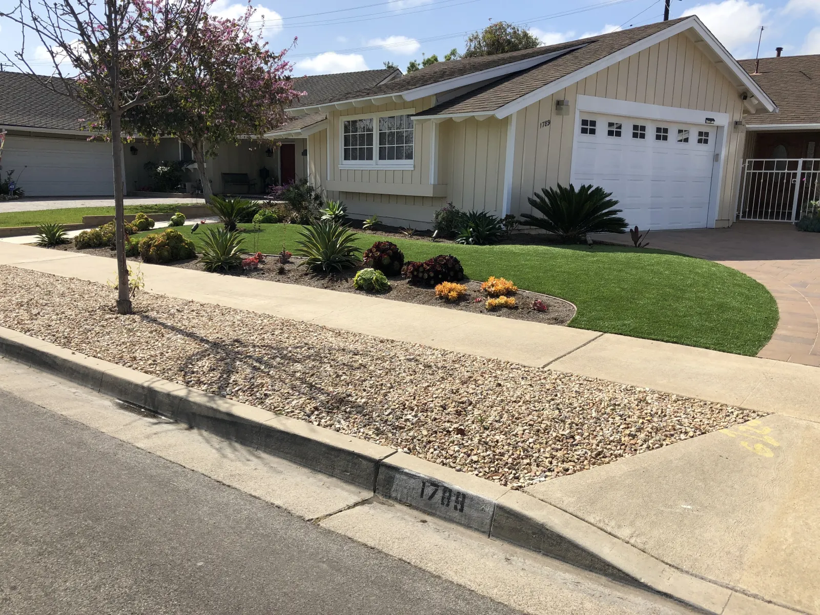 Street view of a suburban house featuring a gravel parking strip, a concrete curb, a green lawn, and a curated front mulch planting bed.