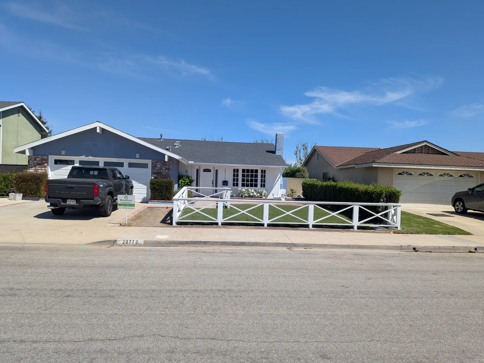 A wide street view of suburban houses, featuring clean driveways and low fences under a blue sky.