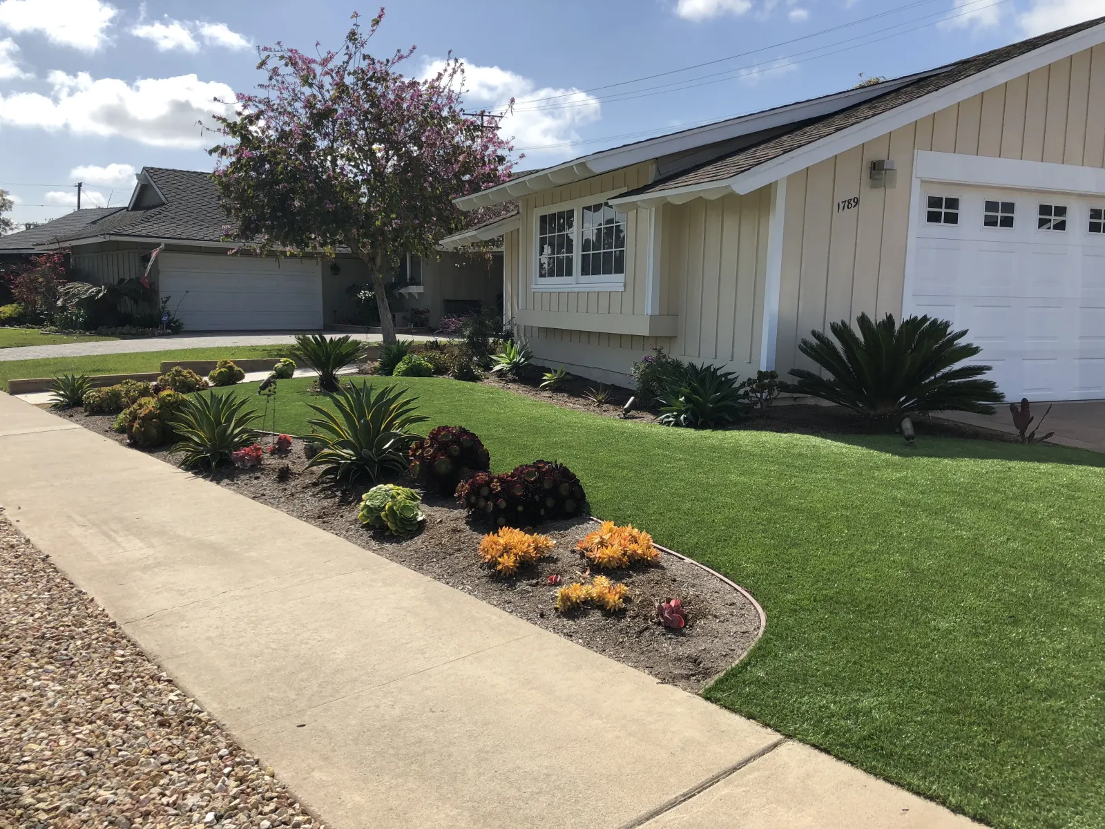Front yard with manicured green grass, a curved planting bed with dark mulch and shrubs, and a concrete walkway leading past a beige house with a garage.