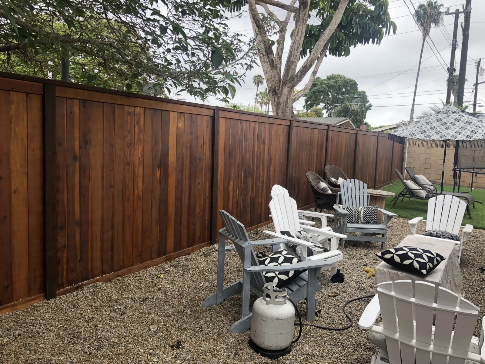 A cozy backyard patio with white plastic armchairs on a gravel base next to a tall wood fence.