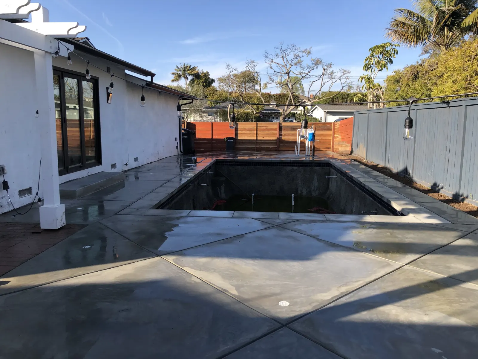 A wide concrete pool deck surrounds an empty swimming pool shell under construction in a residential backyard.