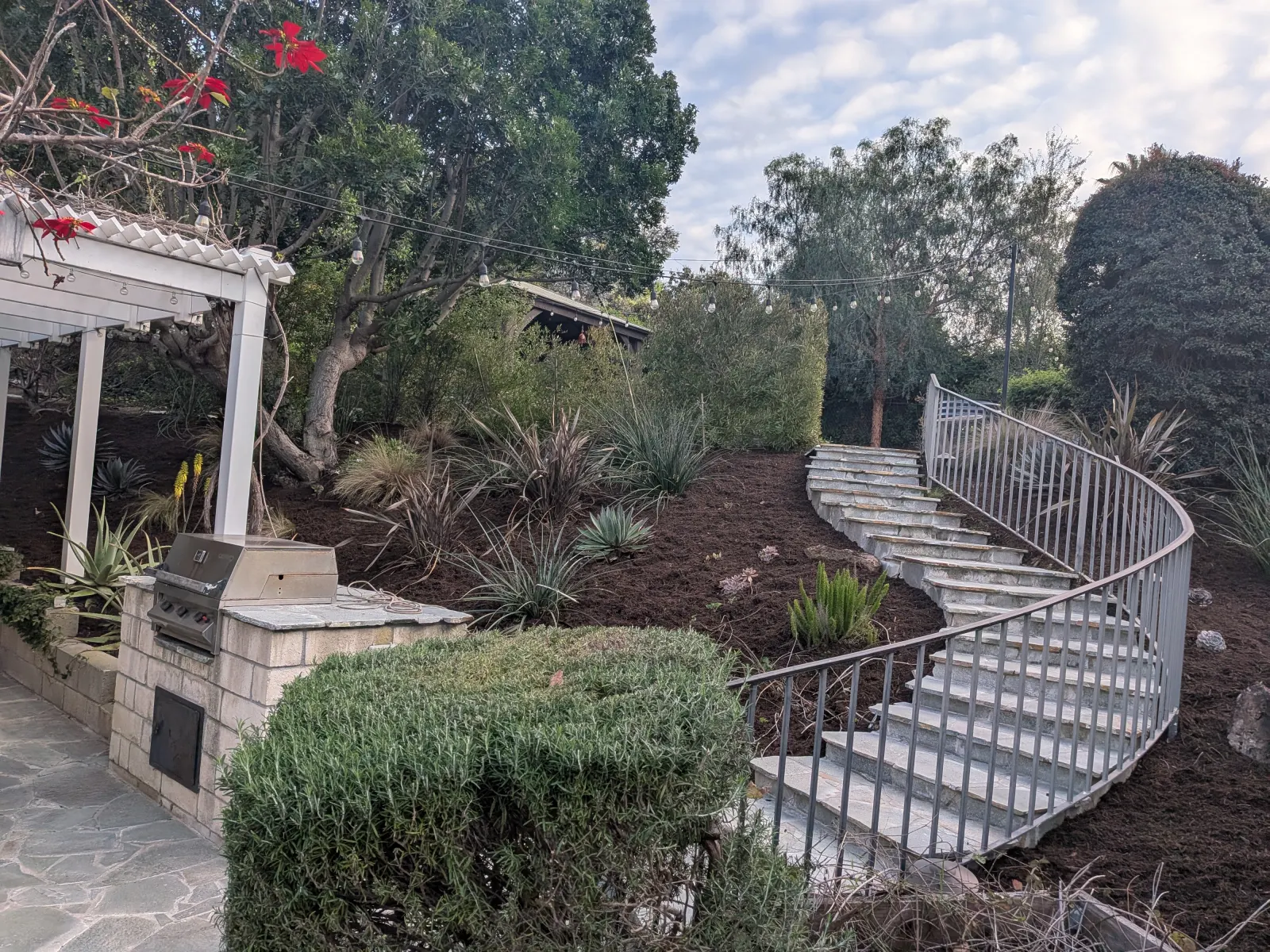 Garden path featuring concrete steps with decorative metal railings leading up a landscaped hill with mulch and green shrubs.