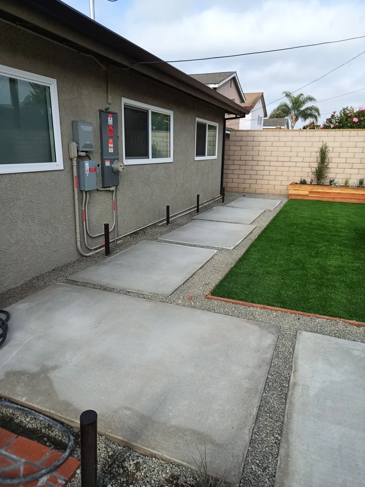 A concrete sidewalk runs along a grey painted house next to artificial turf with decorative borders.