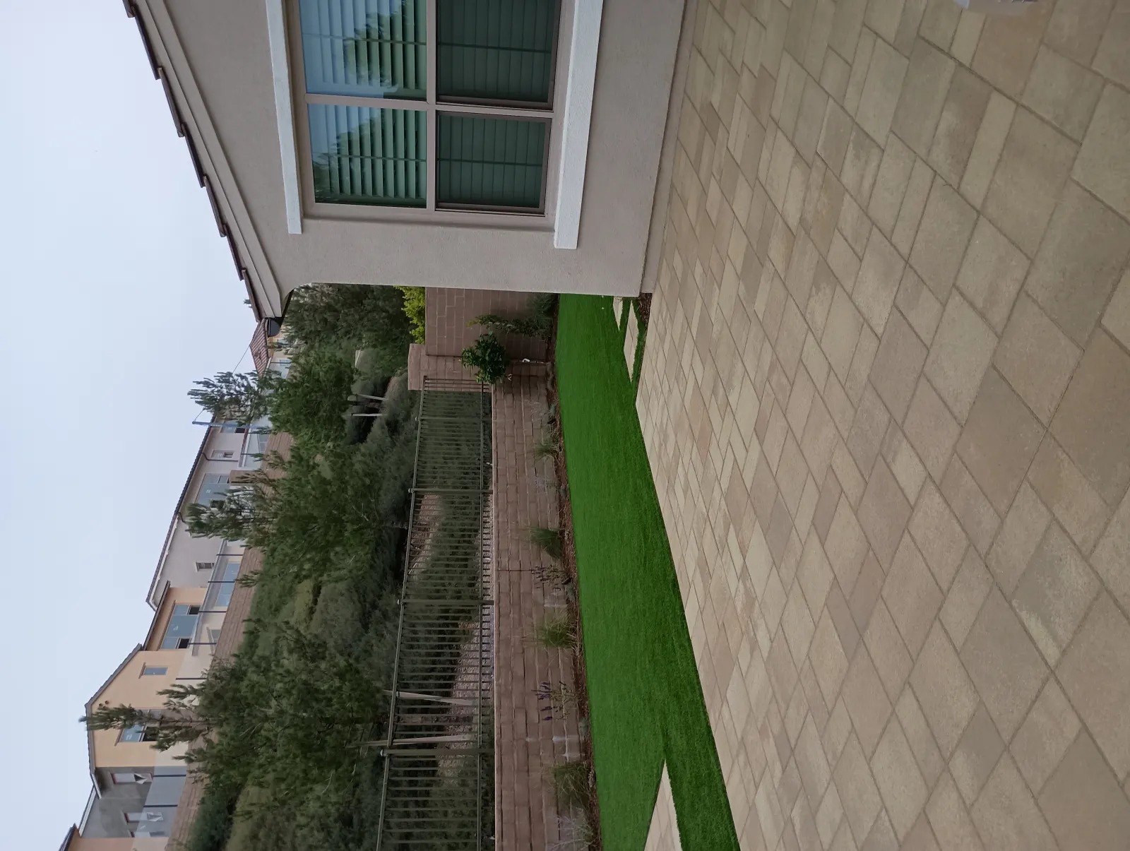 Sideways view of a narrow side yard showing light-colored stone pavers and a strip of bright green artificial turf along a house wall.