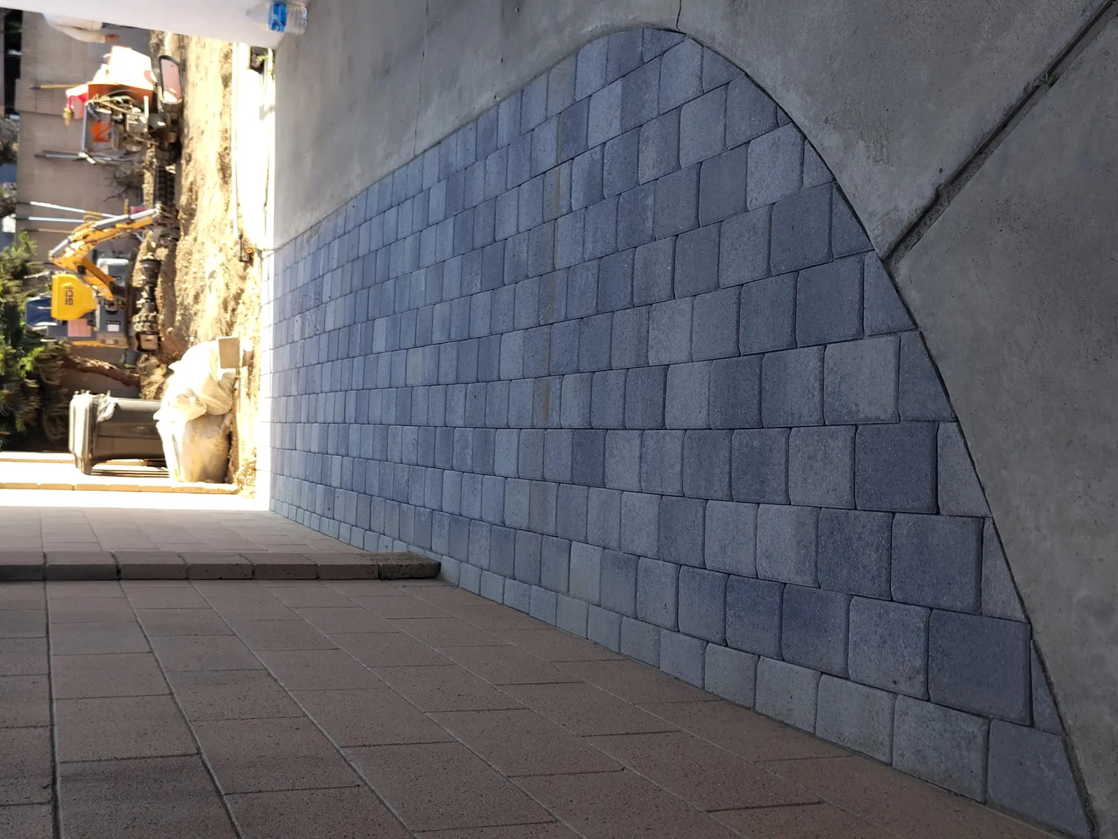 A curved retaining wall made of grey blocks next to steps, with construction equipment in the background.