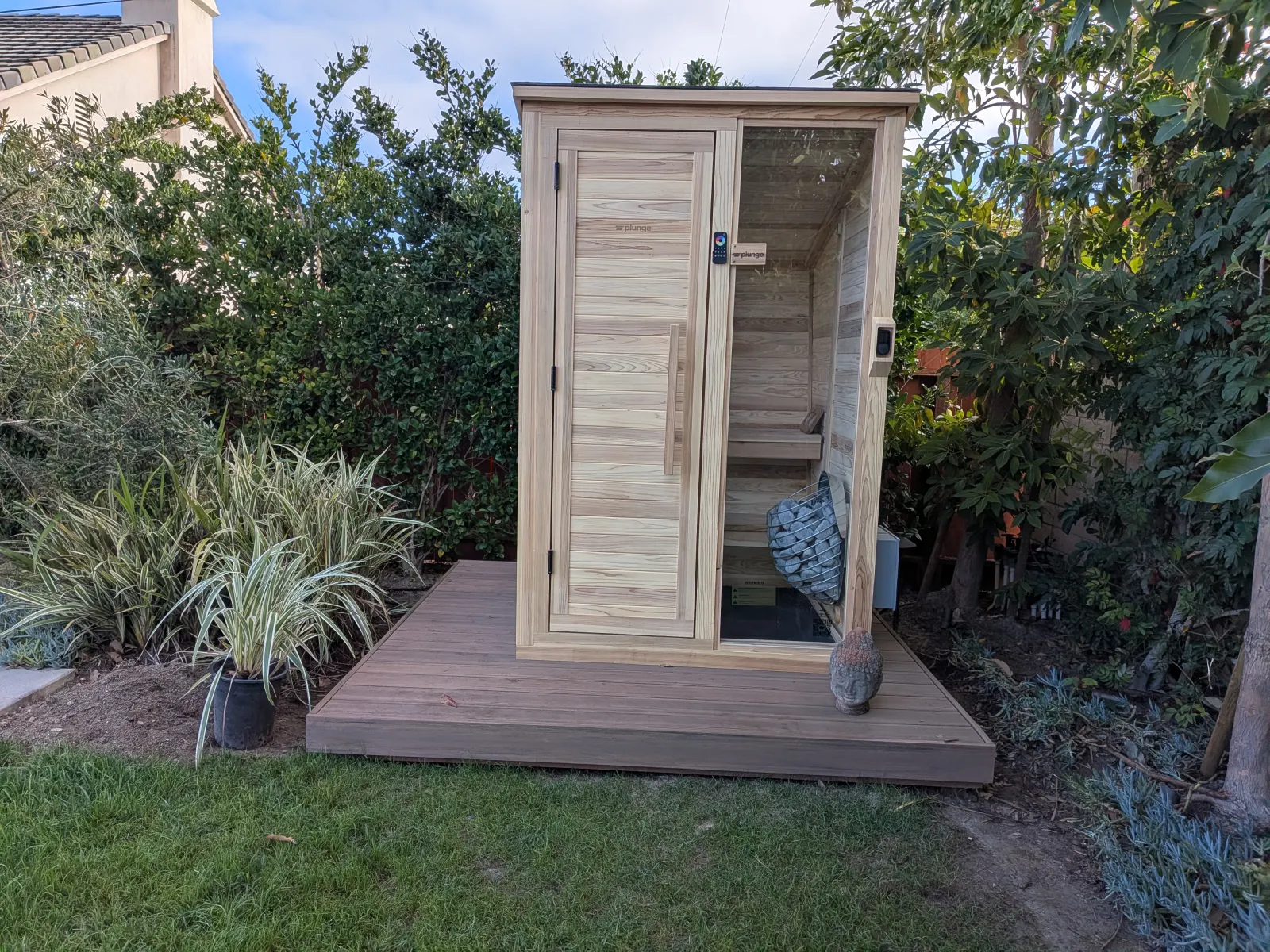 A modern wooden outdoor sauna structure sitting on a small wooden deck platform next to a lawn.