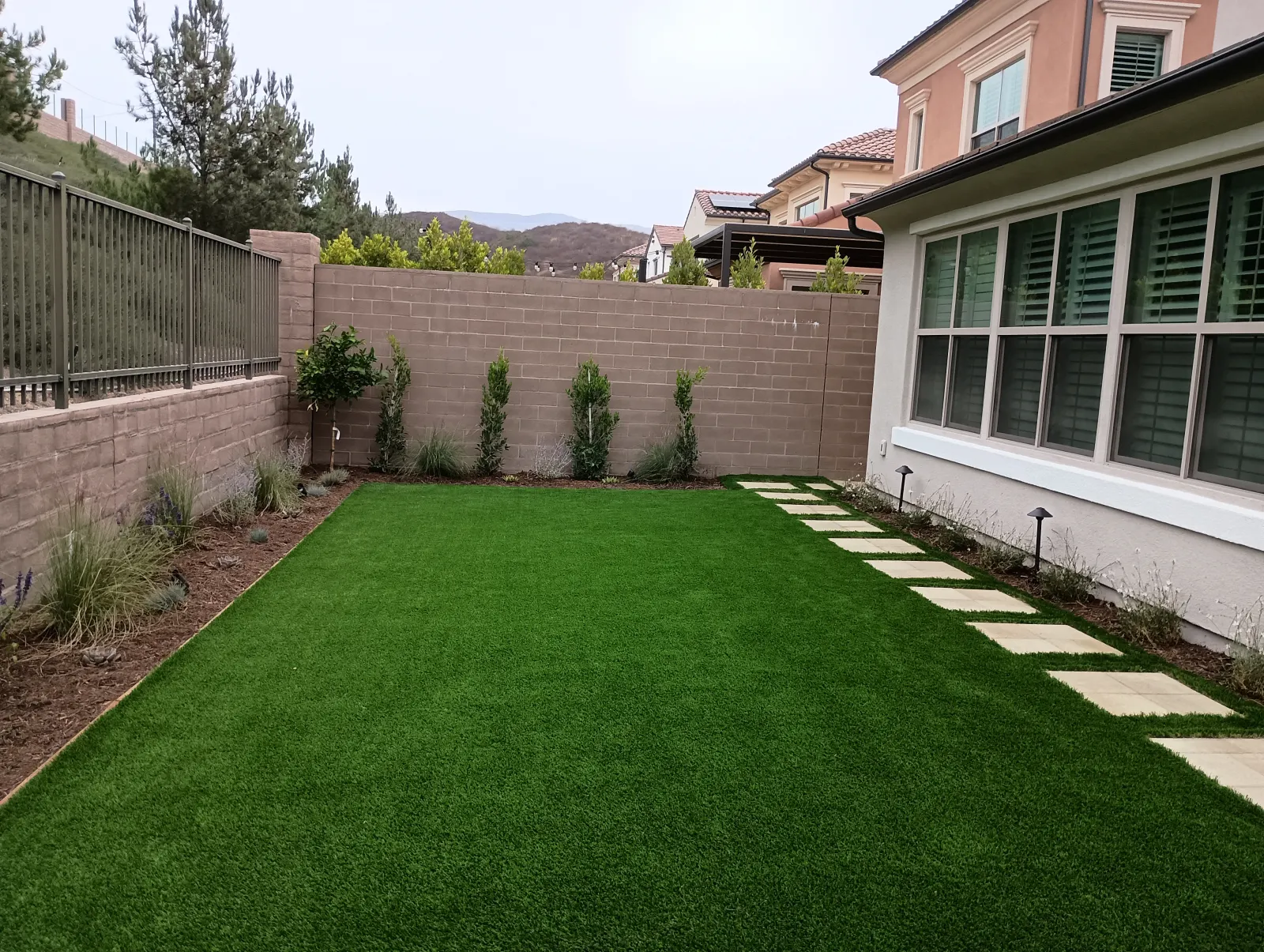 Backyard featuring artificial turf with a path of rectangular light grey stepping stones running along the side of a house.