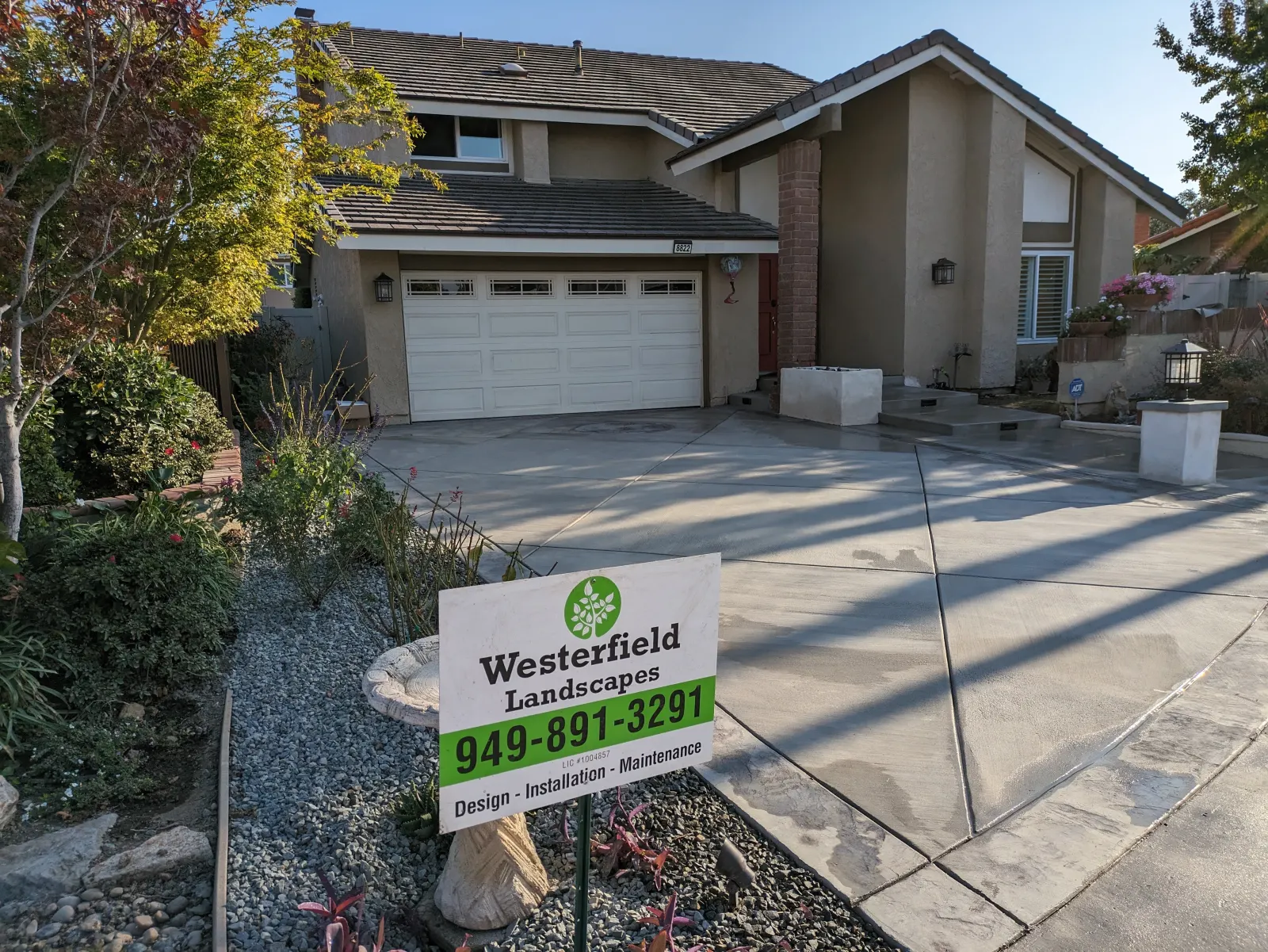 A concrete driveway with geometric joint designs in front of a house, featuring a green landscaper sign.